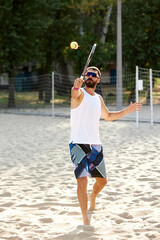 Full-length image of young bearded man in sunglasses playing beach tennis, hitting ball with racket. Summertime outdoor training. Concept of sport, leisure time, active lifestyle, hobby, game, ad