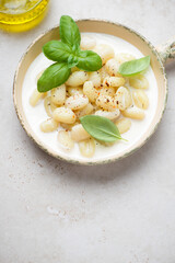 Potato gnocchi with cheese sauce and green basil in a serving pan, vertical shot on a light-beige stone background with space