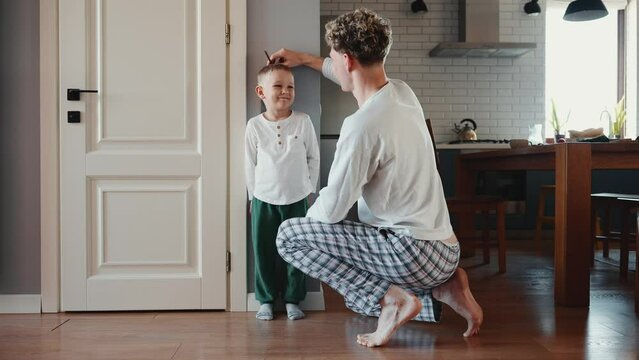 Back view of blond father measures height of his son at home on the wall 
