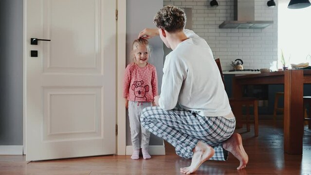Happy blond father measures height of his daughter at home on the wall and then son runs up