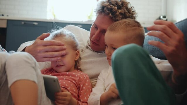 Funny Curly Haired Blond Young Father Watching Cartoons With Kids And Hugging On The Floor At Home