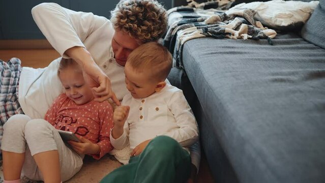 Lovely curly haired blond young father watching cartoons with kids on the floor at home