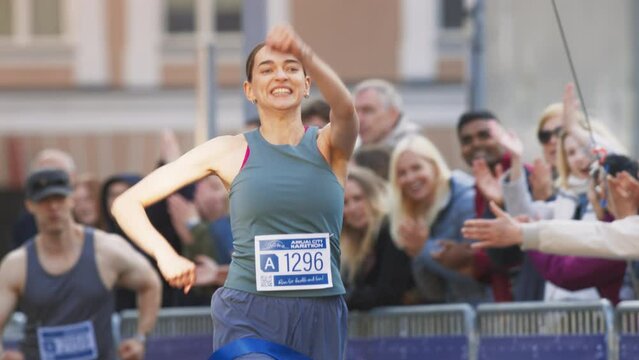 Slow Motion Portrait of Athletic Female Jogger Crossing the Finish Line in Marathon Race with the Audience Cheering. Happy Successful Woman Celebrating Winning, Feeling Empowered with her Achievement