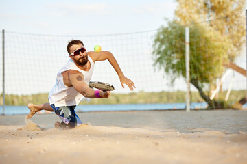 Sportive young man playing beach, paddle tennis, hitting ball with racket and falling down. Outdoor...