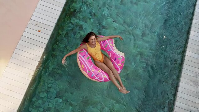 Top view of young woman in swimsuit on the pink donut lilo in the swimming pool. Female relaxing on inflatable float the pool at villa. Inflatable ring and mattress. Summer holiday idyllic. High view