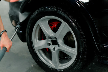 Close-up of a male car wash employee washing the rim and tire of a black luxury car using a high-pressure washer 