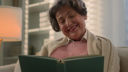 Happy inspired Caucasian old woman reading book smiling nostalgic senior mature lady elderly female retired pensioner grandmother read at home couch watching photos in album memory remember nostalgia