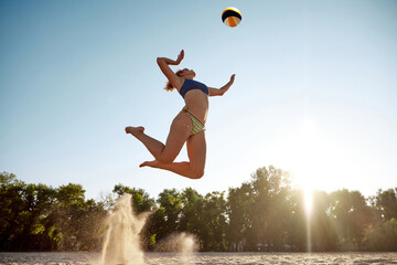 Dynamic bottom view image of young sportive woman playing beach volleyball, hitting ball in a jump...