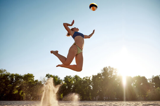 Dynamic bottom view image of young sportive woman playing beach volleyball, hitting ball in a jump over sky background. Concept of sport, active and healthy lifestyle, hobby, summertime, ad