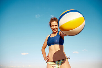 Young smiling woman holding volleyball ball, posing over blue sky background. Outdoor summer training. Concept of sport, active and healthy lifestyle, hobby, summertime, ad