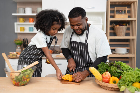 Happy African American kid girl cooking salad with father in kitchen at home