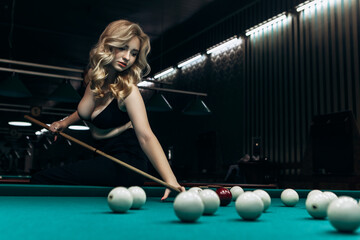 Blond woman playing enjoying billiard, hold cue hit white billiard balls on table with green...