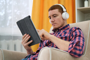 attractive man with freckles in a red plaid shirt with headphones and tablet listening to music at...