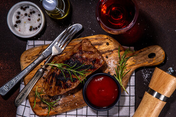 Grilled meat steak on dark table, Pork, veal, lamb, beef steak medium, medium well on cutting board, with spices, ketchup bbq sauce, wine glass. Dinner in steak restaurant menu, top view copy space