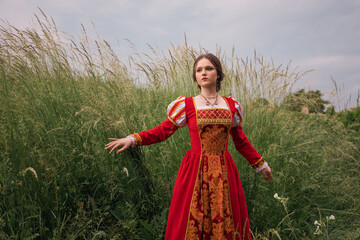 beautiful young woman in a long red medieval dress is standing in the grass in the field under the...