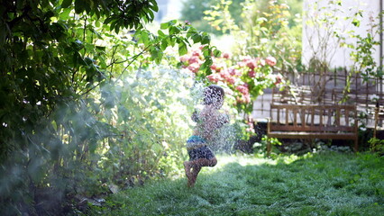 little boy having fun running under water spraying hose in garden during summer day