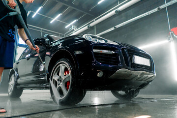 A male car wash employee washes a black luxury car with a high-pressure washer in the car wash bay 