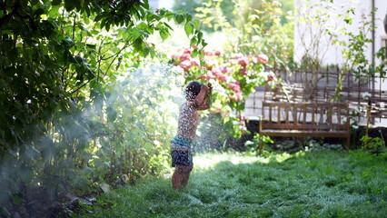 little boy having fun running under water spraying hose in garden during summer day