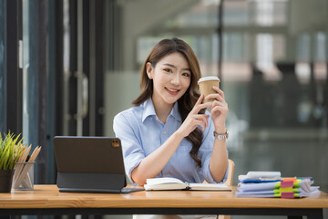 Happy Asian businesswoman working and checking work with a laptop computer while having a cup of coffee at an office.