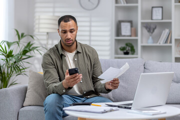 Fototapeta premium African american man working at home with documents, worried sitting on sofa at home and reading letter he received, looking at bills, calling to solve problems