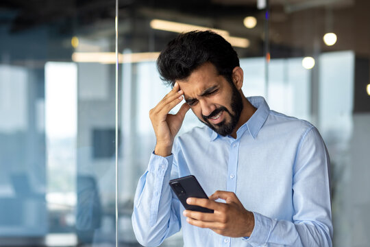 Indian Young Male Businessman Standing In The Office And Looking Upset At The Phone Screen. Holds His Head Anxiously, Received Bad News, Thinks About Solving Problems