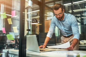 Full concentration. Young bearded businessman in eyeglasses and formal wear examining documents while standing in the modern office