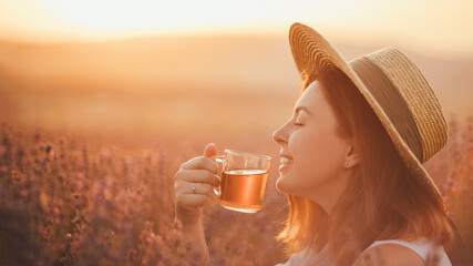 Young happy woman drinking herbal tea, sitting in a beautiful lavender field at sunset.