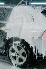 A male car wash employee applies car wash foam to a luxury black car using a spray gun in the car wash box