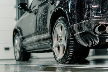 Close-up of car foam application on the rear tire of a black luxury car in the car wash box of the car care concept