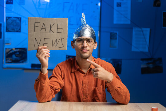 Male conspiracy theorist in a protective foil cap and glasses debunks myths holding poster fake news. Conspiracy theory concept. The schizophrenic works.