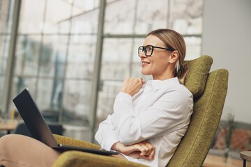 Professional businesswoman sitting in cozy chair and working on laptop on coworking background