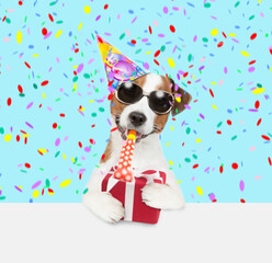 Jack russell terrier puppy wearing sunglasses and party cap blows into party horn and holds big gift box above empty white banner
