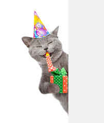Happy cat wearing party cap holds gift box, blows in party horn and looks from behind empty white...