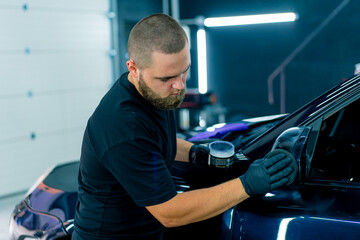 Naklejka na ściany i meble Focused Male car wash worker in black gloves polishes the mirror of a luxury blue car using a polishing sponge car care concept 