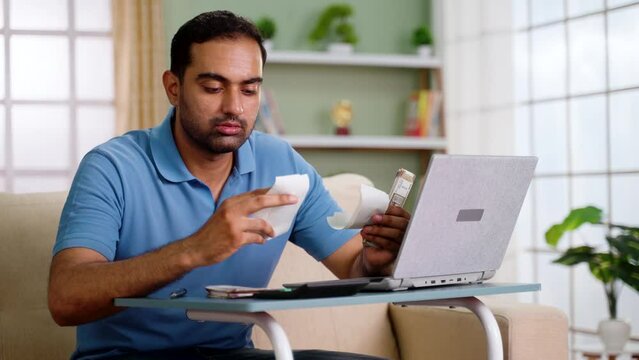 Indian man calculating monthly expenses from bills after salary while sitting on sofa at home - concept of Budget Planning, Money Management and Personal Finance
