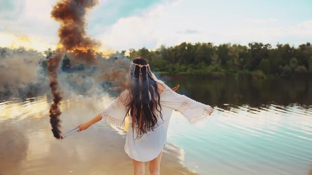 Happy woman dancing spinning waving hands holding smoke bomb colored black color. river bank nature sand beach enjoy life blue sky, long dark hair headband feather boho style. hippie girl white dress 