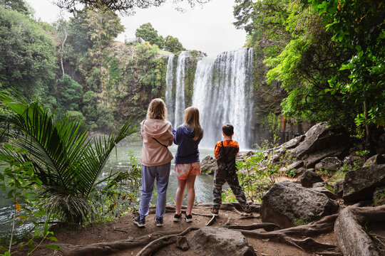 Three people, a mum and two children at an idyllic location looking at a waterfall, with their backs turned. This was taken at Whangarei Falls, New Zealand.
