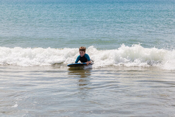 A young boy in a wetsuit is boogie boarding in to shore on a small wave. He is really enjoying playing in the sea. 