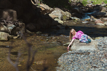 A young girl is reclining next to a small stream with her hand playfully in the water and is truly just enjoying the relaxed moment.