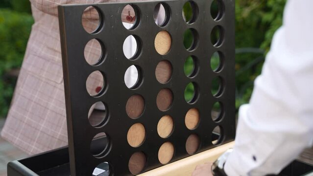Two men in office casual clothes are playing wooden game of four in a row at outdoor party. Confrontation in entertainment at holiday and attempts to win through strategy by throwing mugs into cells.