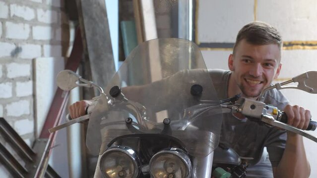 Young man pretending to fast ride a moto in workshop. Cheerful guy fooling around sitting on motorcycle in shed. Happy human enjoying and having fun in garage. Close up Slow motion