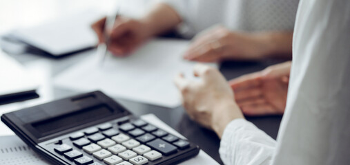 Woman accountant using a pen and laptop computer while counting and discussing taxes with a client, focus on the calculator. Business audit and finance concepts