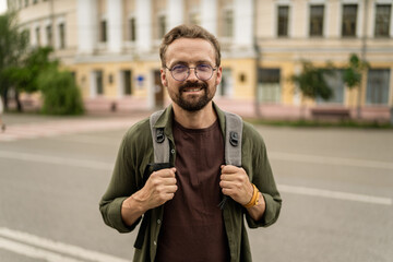 Man wearing glasses and carrying backpack, standing across city. Urban background sets scene for urban adventure, and man's appearance suggests he traveler ready to explore city. . High quality photo