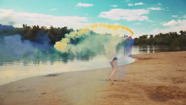 Happy ukranian woman running hands holding smoke bomb blue yellow national colors of flag country Ukraine. river bank enjoy life blue sky, long hair boho style. hippie girl white dress back rear view
