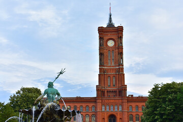 Old building with high clock tower and spire in red brick and a green statue of Neptune in the fountain. Central square. Germany, Berlin, August 2022.