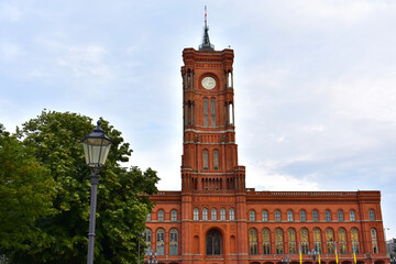 Old building with high clock tower and spire in red brick, green trees and lantern. Central square. Germany, Berlin, August 2022.
