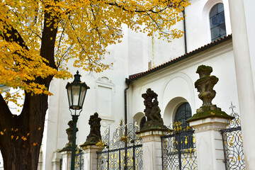 View of the old catholic church with white walls, fence and lantern, and a tree with yellow leaves. Autumn view. Prague, Czech Republic, October 2022.
