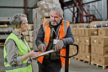 Manager in protective clothing holding documents and giving task to worker during work in warehouse