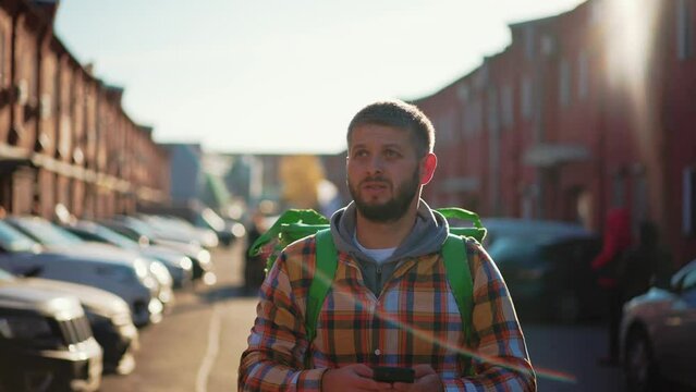 Man courier finding address on map in smartphone on city street looking around. Food delivery guy navigates in phone application with green huge backpack on spine. Fast delivery, courier work concept.