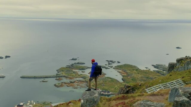 Drone flies aroung young male traveler, standing alone on the stone at the top and enjoying the majestic Lofoten islands landscape
